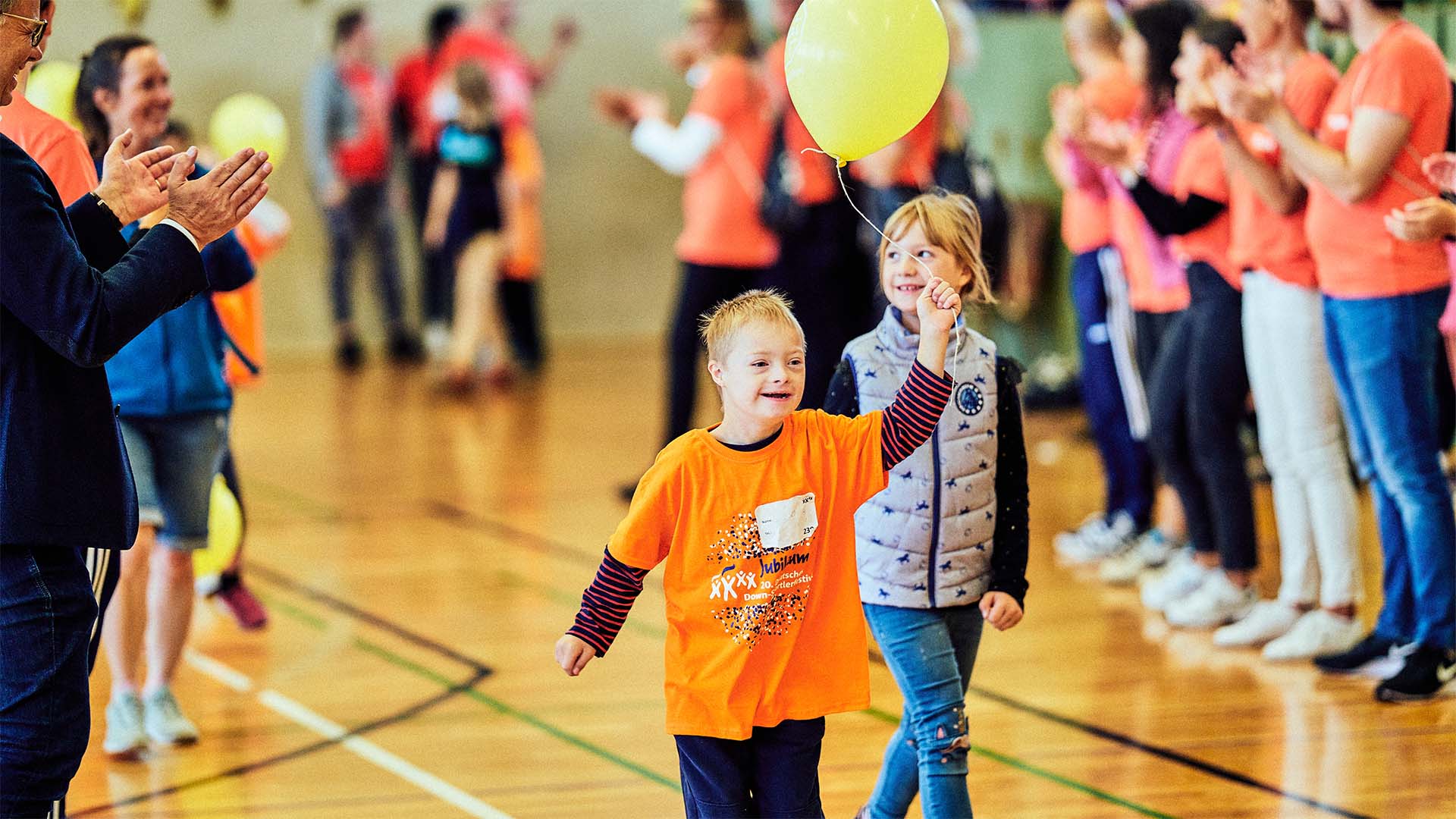 Foto: Zwei Kinder mit gelbem Ballon laufen lachend in einer Sporthalle.