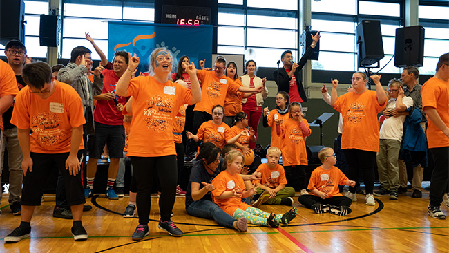 Foto: Gruppe von Teilnehmenden tanzt und jubelt mit erhobenen Händen in einer Halle.