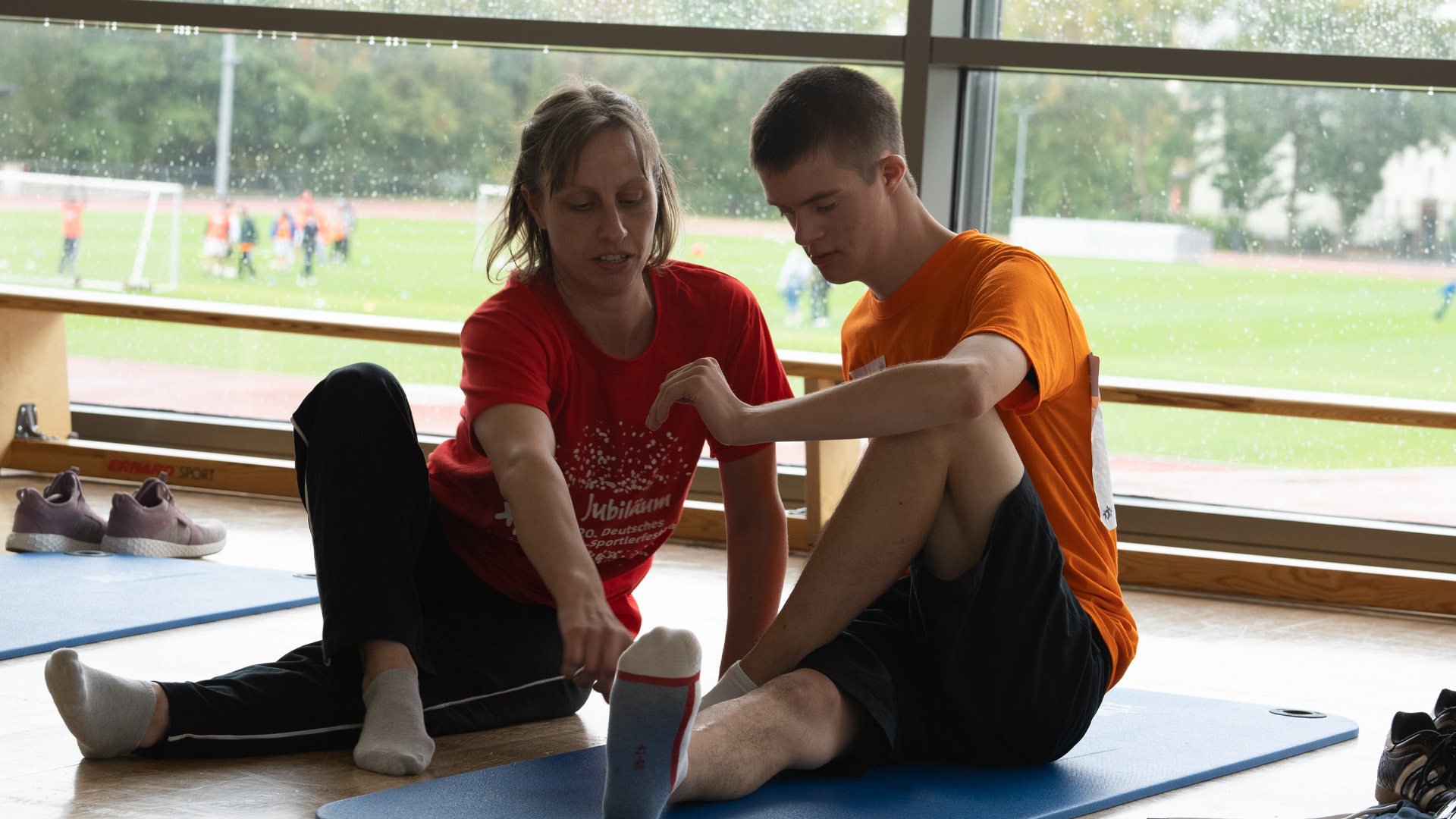 Foto: Frau in rotem Shirt leitet einen jungen Mann mit Down-Syndrom beim Dehnen in der Sporthalle an.
