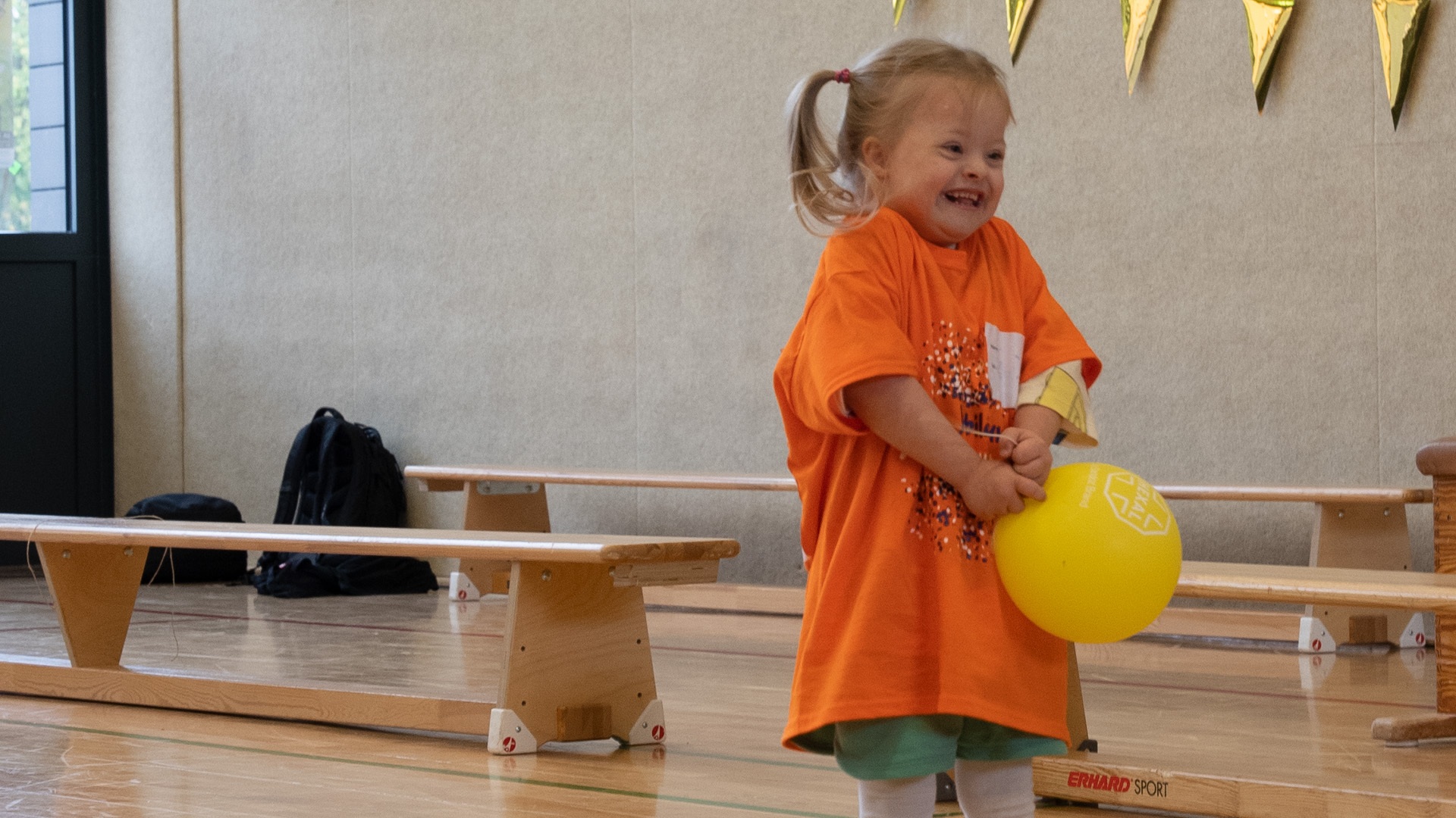 Foto: Lachendes kleines Mädchen in der Turnhalle hält einen gelben Ballon in den Händen.