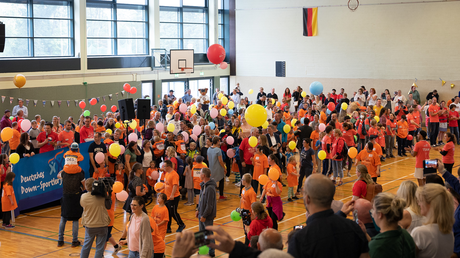 Foto: Große Gruppe von Teilnehmern und Publikum feiert und applaudiert in der vollen Sporthalle.