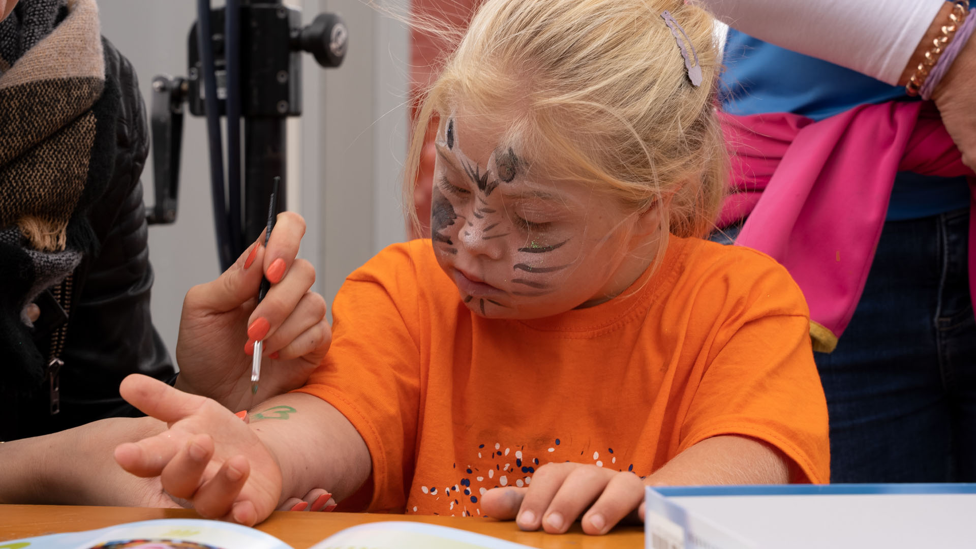 Foto: Mädchen mit bemaltem Gesicht schaut zu, wie ihr Handgelenk bemalt wird.