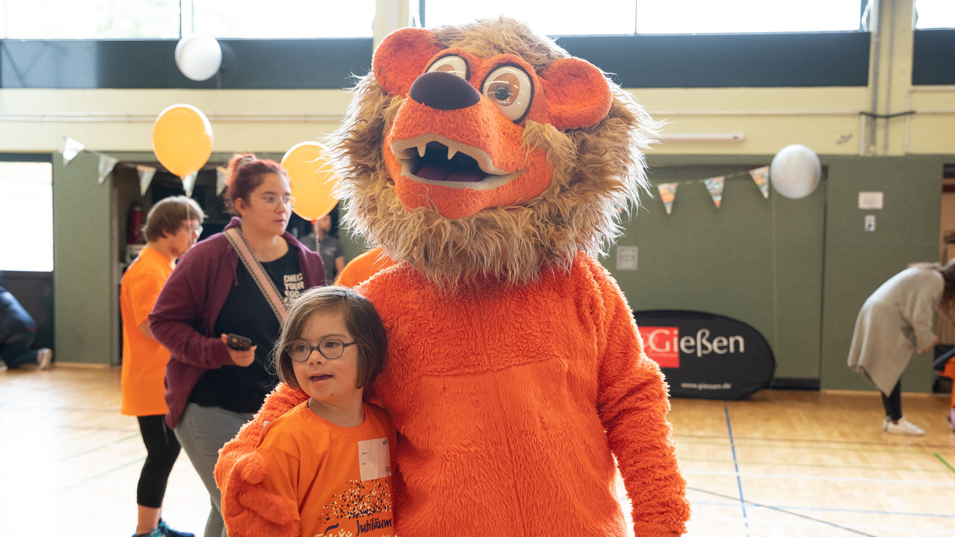 Foto: Lachendes Mädchen im orangenen Shirt umarmt das orangefarbene Löwen-Maskottchen.