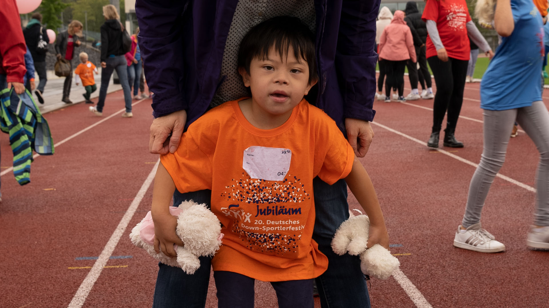 Foto: Kleines Kind mit Down-Syndrom hält zwei Stofftiere und steht vor einer Begleitperson