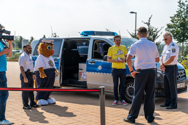 Foto:  Moderator André Gatzke steht Maskottchen neben einem Polizeiauto, umgeben von Polizisten.