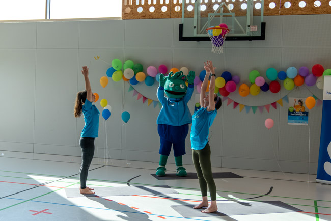 Foto: Yoga-Trainerinnen und Maskottchen führen Dehnübungen mit erhobenen Armen durch.