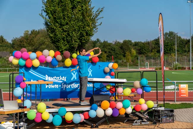Foto: Trainer auf Bühne macht Aufwärmübungen mit erhobenen und gedehnten Armen.