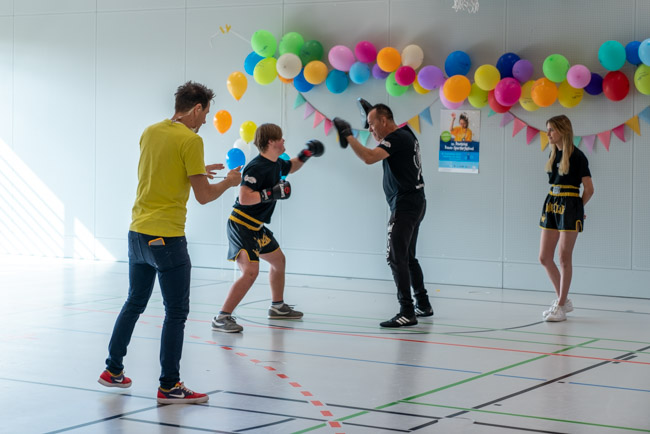 Foto: Jugendlicher im Box-Outfit trainiert mit Trainer in der Halle, Moderatoren beobachten.