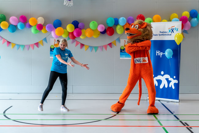 Foto: Trainerin tanzt mit dem orangenen Löwen-Maskottchen in der bunt geschmückten Halle.