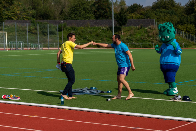 Foto: Zwei Männer geben sich auf dem Rasen einen Faustgruß, während das Maskottchen zuschaut.