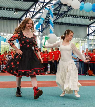 Foto: Model Tamara Röske und ein Mädchen, beide in Kleidern tanzen, vor Zuschauern in der Halle.