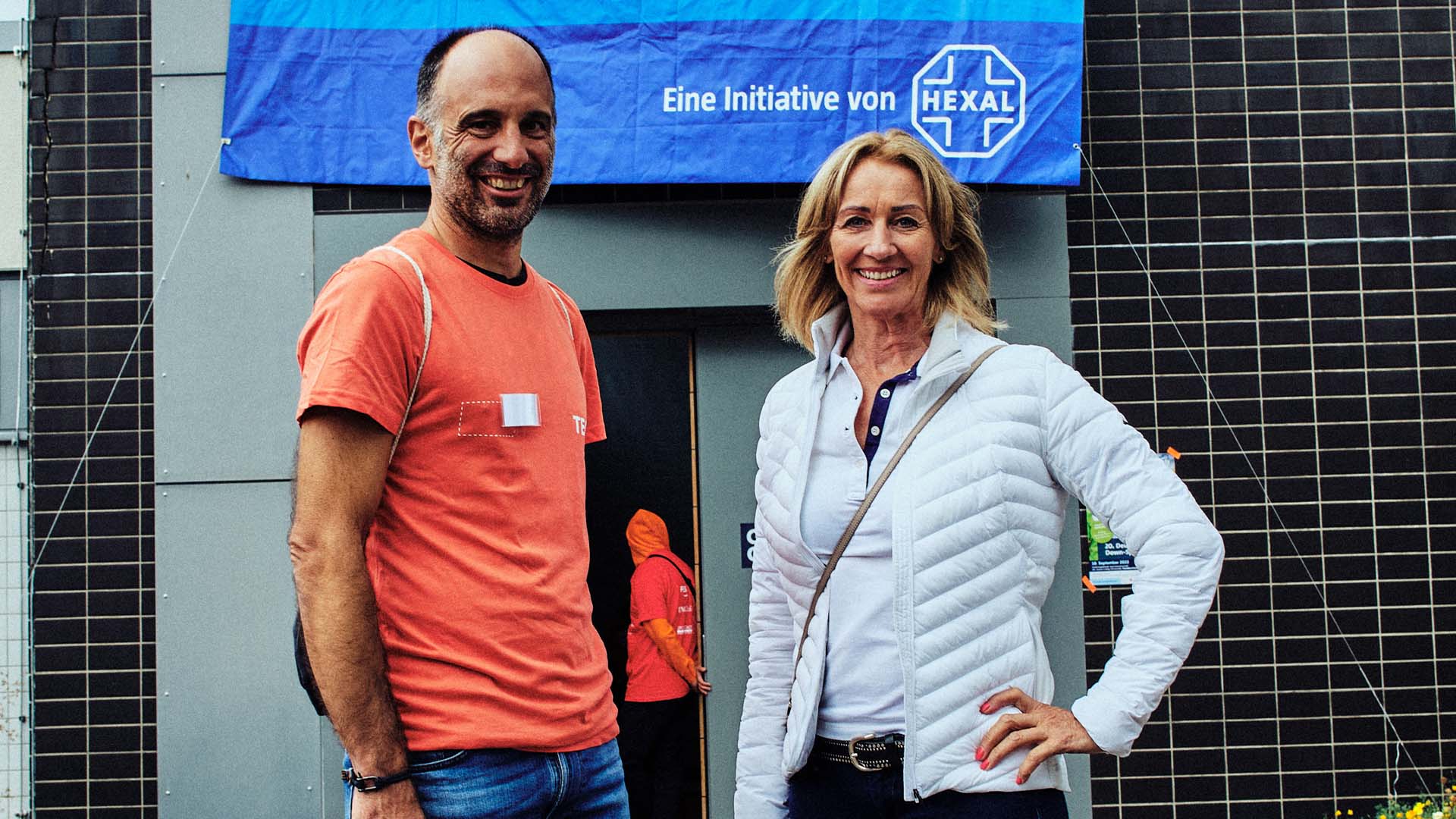 Foto: Lächelnder Mann im orangenen Shirt und lächelnde Frau posieren vor blauem Event-Banner.