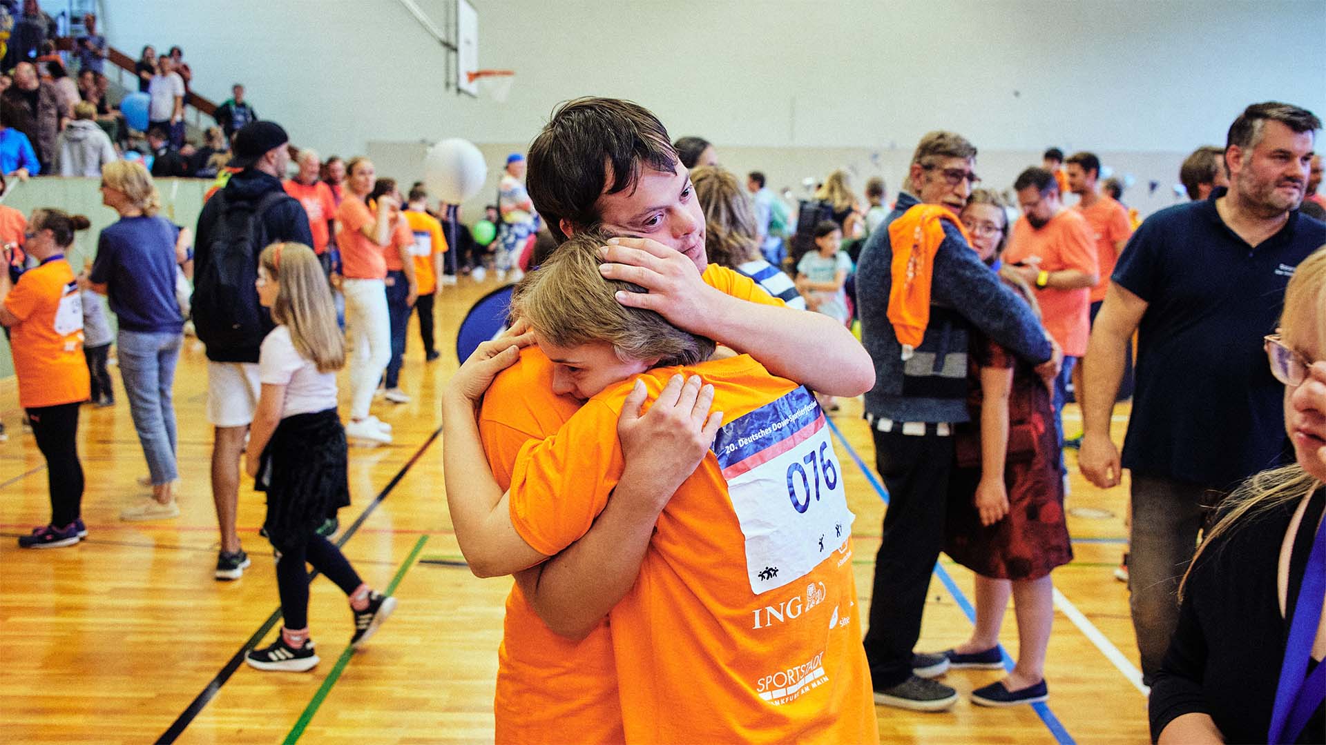 Foto: Zwei Teilnehmer in orangenen Shirts umarmen sich in einer Sporthalle.