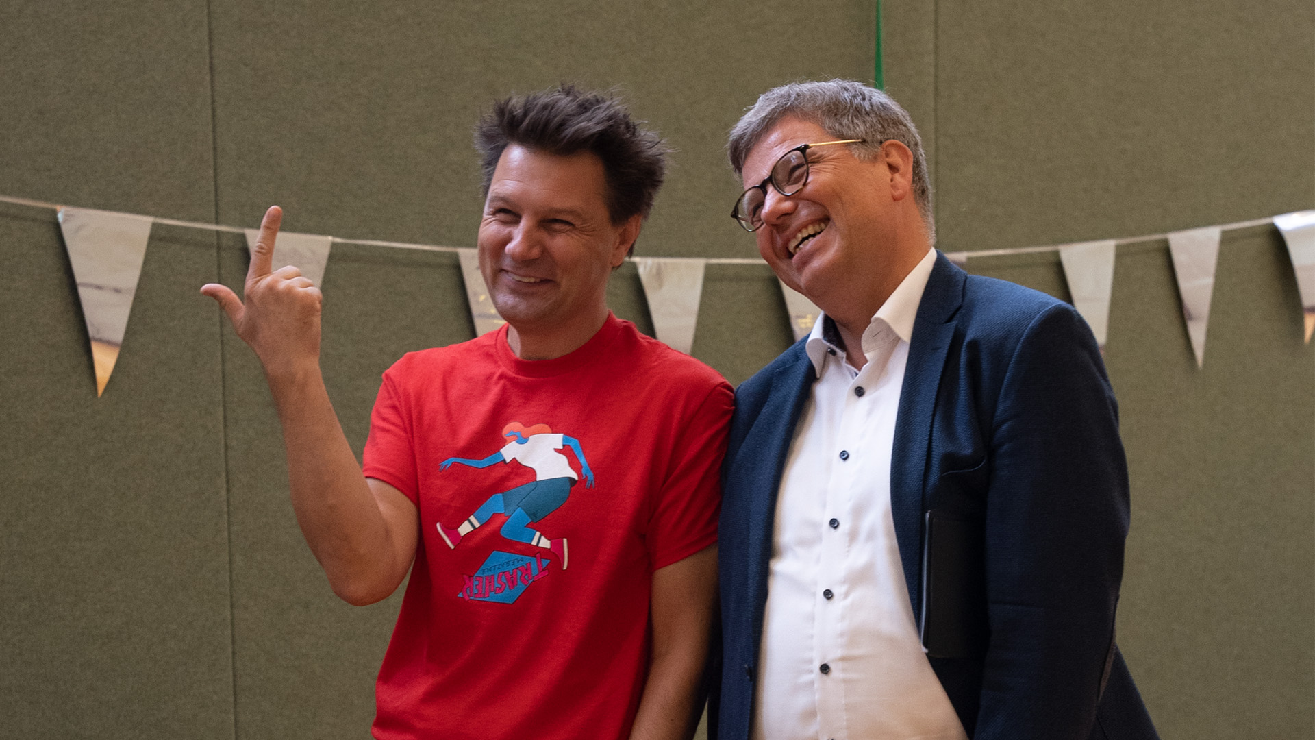 Foto: Moderator André Gatzke hebt den Zeigefinger, während ein Mann im Anzug ihm zulächelt.