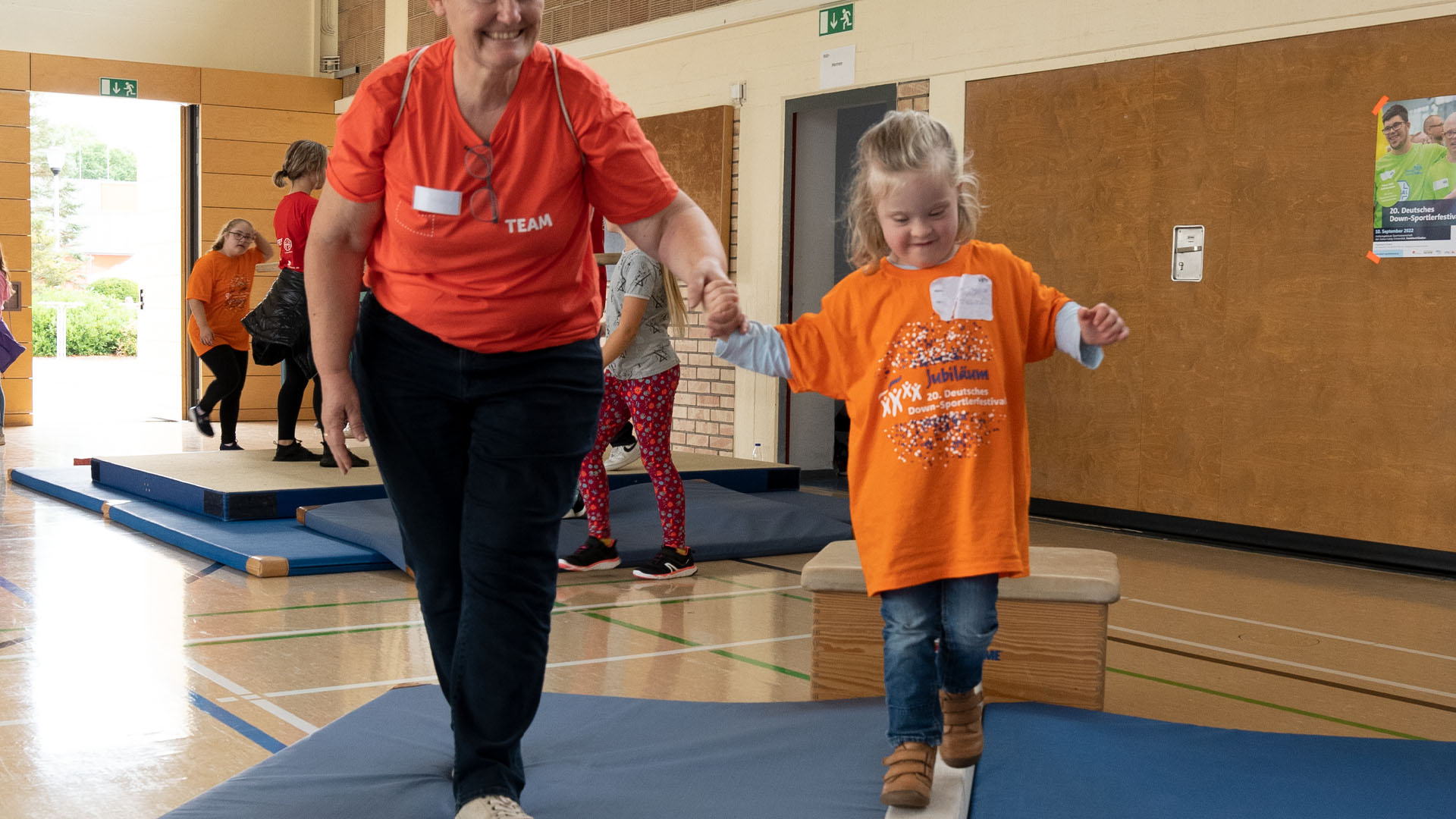 Foto: Erwachsene Frau hält ein Mädchen an der Hand, das auf einem schmalen Holzbalken balanciert.