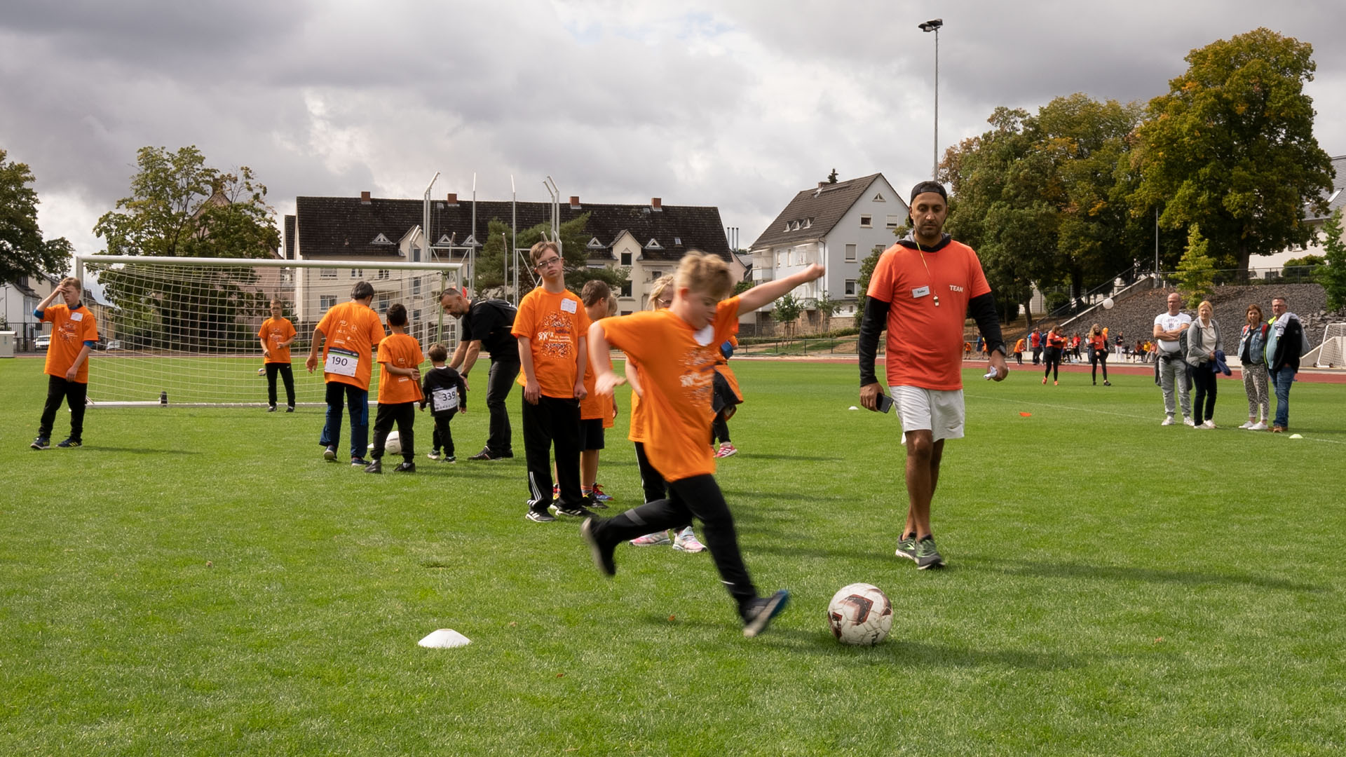 Foto: Kind im orangenen Shirt schießt auf dem Rasen beim Fußballtraining den Ball.